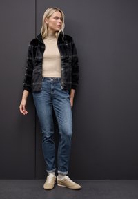 Chaqueta de pelo sintético negra, suéter de cuello alto beige, jeans azules y zapatillas beige. La chaqueta cuenta con un cierre de cremallera y puños en contraste.