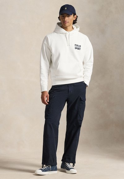 Fehér kapucnis pulóver fekete "POLO SPORT" felirattal, sötétkék cargo nadrág, és kék sportcipők fehér talppal. Kényelmes, laza szabású hétköznapi öltözék.