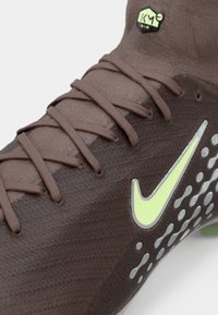 Κοντινή λήψη καφέ αθλητικού παπουτσιού με σφιχτά δεμένα κορδόνια, νέον κίτρινο λογότυπο Nike και λογότυπο "KM AIR" στον γιακά που μοιάζει με κάλτσα.