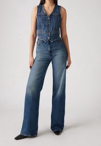 Femme portant un gilet en denim sans manches boutonné et un jean taille haute à jambes larges avec des chaussures noires pointues, debout sur un fond clair.
