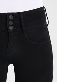 Vaqueros de mezclilla negra con una cintura alta, cierre de tres botones y un bolsillo lateral con un sutil detalle de costura curva.