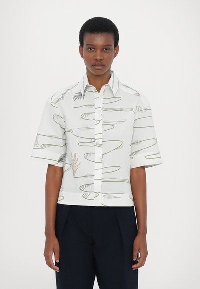 Kurzärmliges, cropped weißes Shirt mit wellenförmigen grünen und orangenen Linienmustern. Knopfleiste vorne und klassischer Kragen. Glatte Stoffoberfläche.