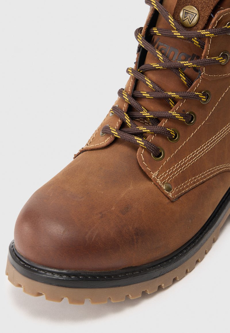 Brązowy skórzany but sznurowany z żółto-paskowanymi brązowymi sznurówkami, metalowymi oczkami, haftowanymi detalami i gumową podeszwą z bieżnikiem.