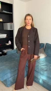 Hnědý texturovaný kardigan s knoflíky, kombinovaný s hnědými širokými kalhotami. Drží hnědé psaníčko, stojí před modrou látkovou pohovkou.