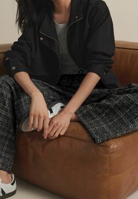 Veste noire avec fermeture éclair dorée, haut gris et pantalon large à motifs. Le modèle porte des chaussures blanches et noires, assis sur un pouf en cuir marron.