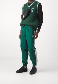 Grön ribbad stickad tröja med vita detaljer, kombinerad med gröna track pants med tre vita ränder och svarta sneakers.