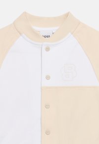 Λευκό και μπεζ μακρυμάνικο πουκάμισο με μπεζ κουμπιά τύπου snap και ένα διακριτικό ανάγλυφο λογότυπο "B" στο στήθος.