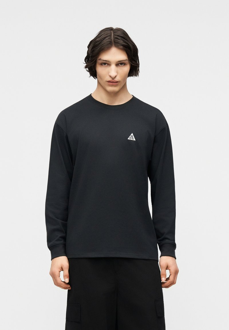 Νεαρό άτομο με μεσαίου μήκους σκούρα μαλλιά φοράει μαύρο πουκάμισο με μακριά μανίκια και μικρό λευκό τρίγωνο λογότυπο στο στήθος.