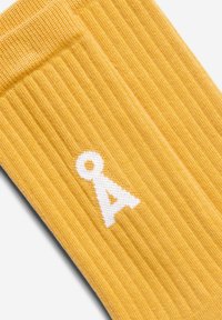 Gelber, gerippter Socken mit einem weißen „A“-Logo. Das Material wirkt weich mit einer strukturierten Oberfläche. Entworfen für Komfort und Stil.