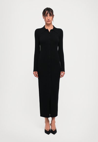 Donna in piedi con un vestito aderente lungo nero a costine, con colletto e maniche lunghe, che indossa tacchi a punta neri.