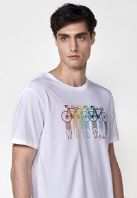 T-shirt bianca in cotone con una grafica di bicicletta colorata dalle tonalità arcobaleno e linee verticali, accompagnata dal testo "KOROSHI" stampato sotto.