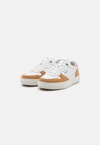 Bílé tenisky s hnědými semišovými akcenty, se perforovanou špičkou, plochými tkaničkami a bílou gumovou podešví. Logo na boku a na jazyku.