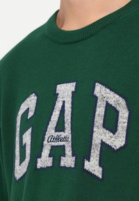 Grüner Pullover mit großem, in Weiß gesticktem "GAP"-Logo und blauer Umrandung, strukturiertem Stoff und geripptem Rundhalsausschnitt.