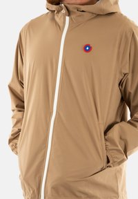 Veste légère couleur marron clair avec une fermeture éclair blanche, capuche ajustable et un logo rond bleu et rouge sur la poitrine. Elle présente des poignets élastiques et des poches latérales.