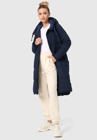 Navy long puffer coat with hood, quilted texture, front zipper closure, and side pockets, worn over cream joggers and light sneakers.