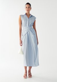 Hele helesinine midi kleit, millel on krae, ilma varrukateta disain, sõlmitud vöökoht ja kaks esikotti. Aksessoriseeritud väikese valge käekotiga.