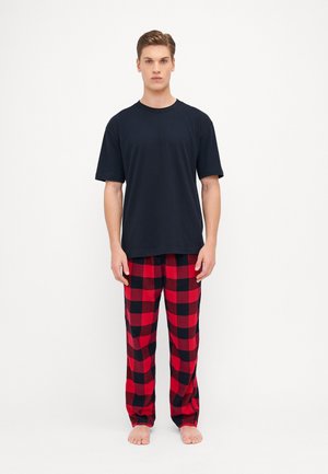 Outfit da uomo composto da una maglietta nera in cotone e pantaloni da pigiama in flanella a quadri rossi con quadrati neri e rossi. Posizione a piedi nudi, sfondo neutro.