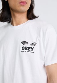 Bela bomba majica iz bombaža z grafičnim potiskom oči nad napisom "OBEY HOUSE OF SUBVERSION" v krepki črni pisavi. Okrugli izrez.