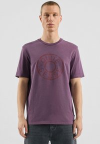 Fioletowy bawełniany t-shirt z krótkimi rękawami i okrąg�łym dekoltem, z dużym, okrągłym, wyblakłym napisem „QUITE SENSE” w kontrastującym czerwonym kolorze.