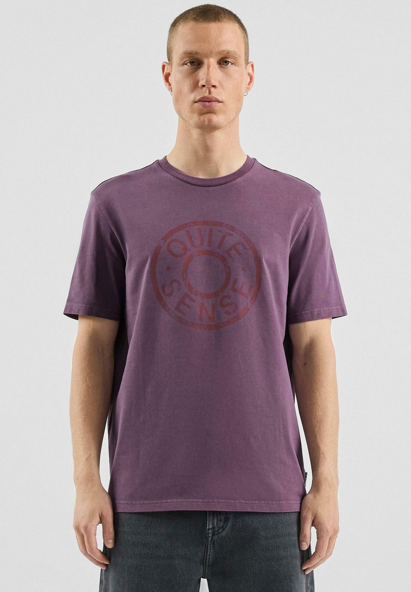 Fioletowy bawełniany t-shirt z krótkimi rękawami i okrągłym dekoltem, z dużym, okrągłym, wyblakłym napisem „QUITE SENSE” w kontrastującym czerwonym kolorze.