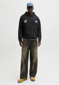 Sudadera negra con logo blanco, de corte relajado. Jeans de pierna ancha desgastados con acabado desvaído. Gorra azul clara. Calzado amarillo con suela texturizada.