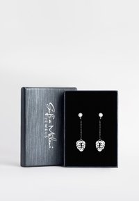 Silverfärgade hängande örhängen med bladformade hängen på kedjor, placerade i en svart sammetdisplaylåda med en texturerad silveryta.