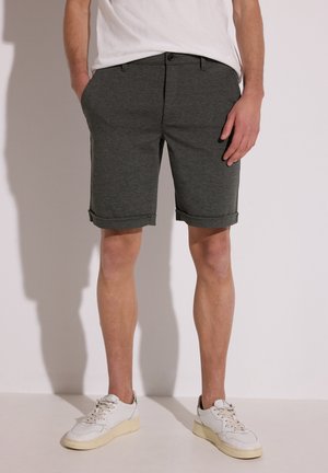 Mann trägt dunkelgraue Shorts mit umgeschlagenem Saum und weiße Sneakers, steht mit den Händen in den Taschen vor einem einfarbigen Hintergrund.