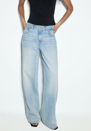 Person iført lyseblå vidde jeans og en sort ærmeløs top, står med hænderne i jeanslommerne mod en ensfarvet hvid baggrund.