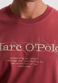 Rode katoenen t-shirt met geborduurde tekst in crèmewit, voorzien van stiklijnen en een slogan over natuurlijk erfgoed en bescherming van het landschap.