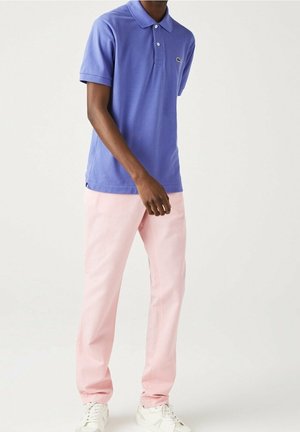 Paarse poloshirt met een kraag en drie knopen, gecombineerd met lichtroze broek en witte sneakers. Lacoste-logo op het shirt.