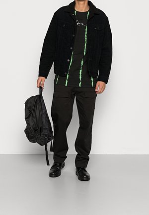 Zwarte corduroy jas met een fuzzy kraag over een zwart tie-dye shirt met groene verticale strepen, gecombineerd met zwarte cargo broek en schoenen.