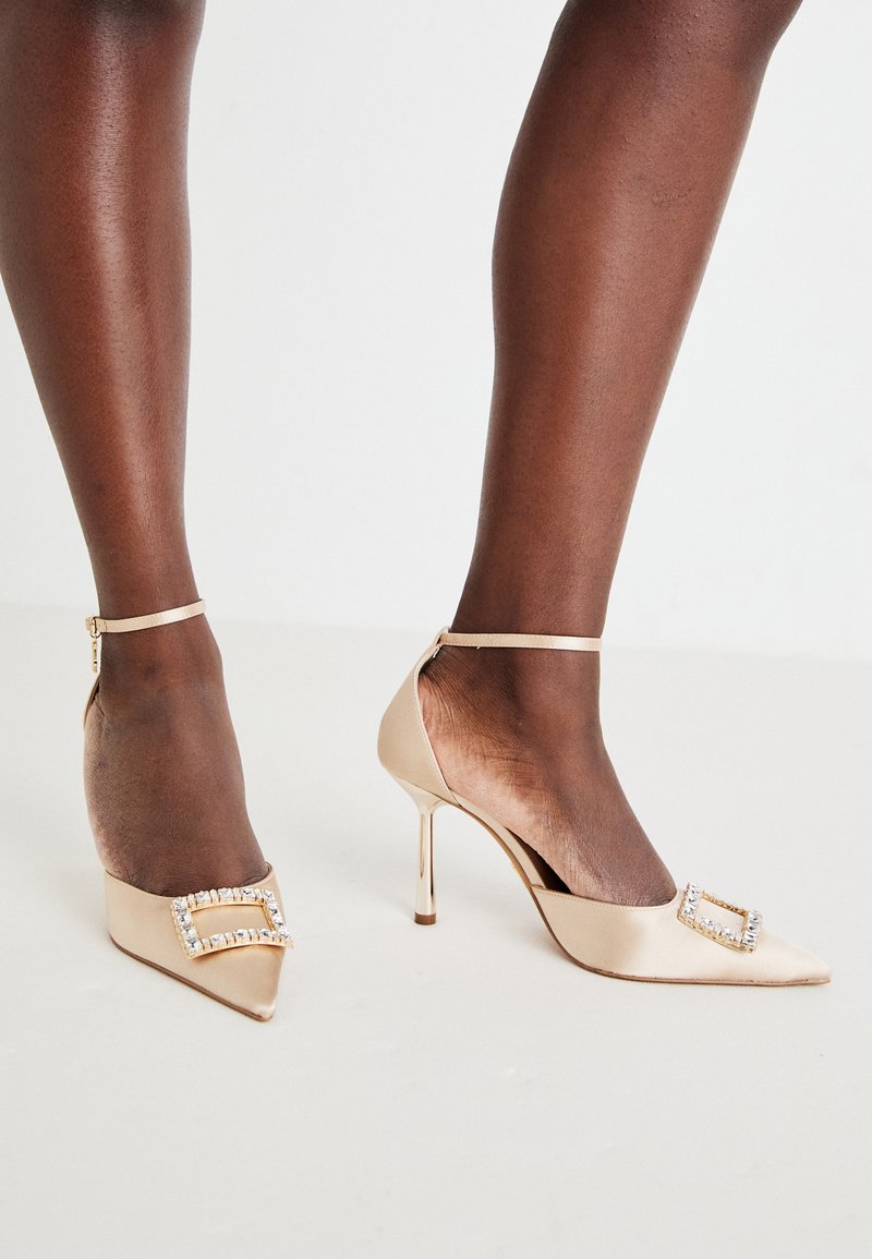 Beige Satin High Heels mit spitz zulaufenden Zehen, ausgestattet mit einem mit Juwelen verzierten Schnallen-Detail und einem Knöchelriemen. Glänzende Stiletto-Absätze.