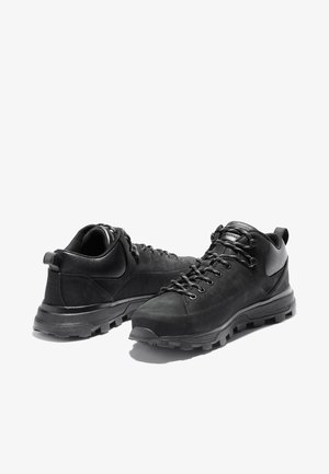 Paire de chaussures de randonnée noires avec semelles à crampons épaisses, cols rembourrés et lacets à l'avant sur fond blanc.