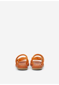 Une paire de sandales orange avec des brides à la cheville et des boucles, des semelles plates tissées, présentée de l'arrière sur un fond blanc.