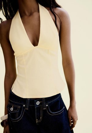 Femme portant un top à col halter jaune pâle avec un profond décolleté en V et un jean en denim bleu foncé, debout devant un fond neutre clair.