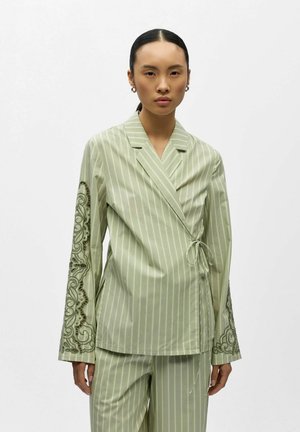 Vrouw draagt lichtgroene, gestreepte wikkelblazer en bijpassende broek, met geborduurde details op de mouwen, staand tegen een witte achtergrond.