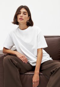 White cotton t-shirt with short sleeves, round neckline, relaxed fit. Brown wide-leg trousers, seated on a brown leather couch.