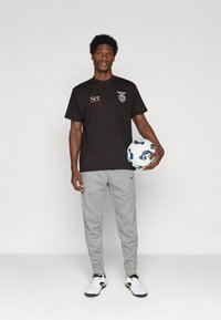 T-shirt preto de manga curta com logos, calças de moletom cinzentas, sapatos desportivos brancos, e uma bola de futebol azul e branca, em pé contra um fundo neutro.