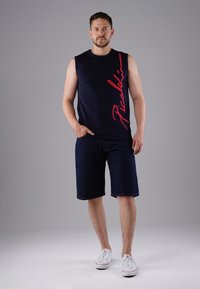 Navy sleeveless t-shirt with red "Picabia" print, paired with dark blue denim shorts and white sneakers. Smooth fabric, relaxed fit.