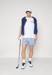Hvid t-shirt, marineblå håndklæde, mønstrede blå shorts med cirkulært design, hvide sko, holder en golfkølle, iført en hvid kasket og handske.