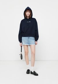 Kvinne iført en marineblå hettegenser med tekst, lys blå denim miniskjørt, hvite sokker, svarte loafers, holder en liten mønstret håndveske ved siden av seg.