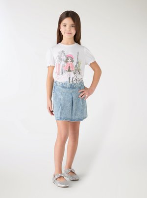 Ragazza che indossa una maglietta bianca a tema Parigi, gonna di jeans scintillante e scarpe Mary Jane glitter argento, in piedi con una mano sui fianchi.