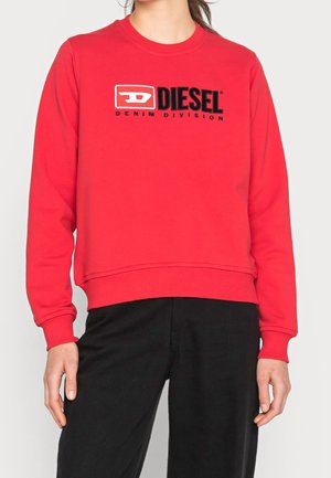 Personne portant un sweat-shirt rouge Diesel avec logo et texte "Denim Division" associé à un pantalon noir sur un fond uni.