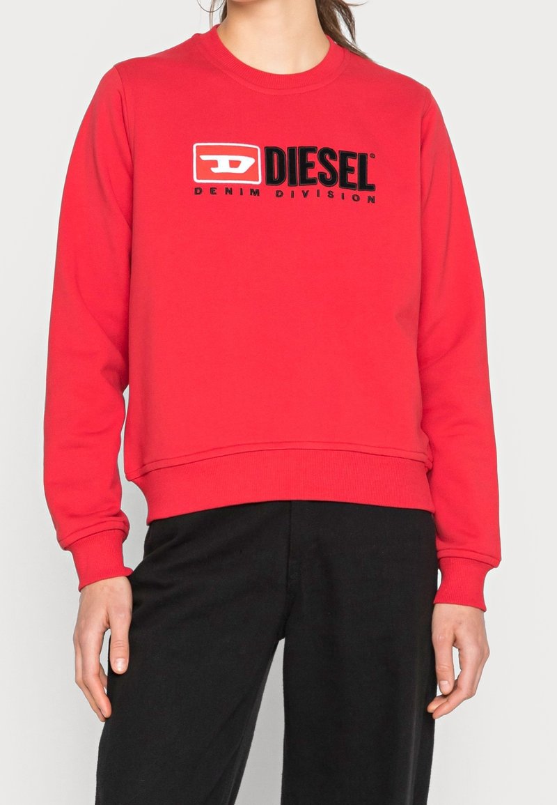 Personne portant un sweat-shirt rouge Diesel avec logo et texte "Denim Division" associé à un pantalon noir sur un fond uni.