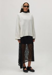 Witte gebreide trui met ribbelmanchetten, gedragen over een zwarte kanten maxi-jurk. Zwarte zonnebril en schoenen maken de look af.