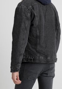 Black denim jacket with a classic design, featuring a collared neck, button closures, and a faded texture. Worn over dark jeans.
