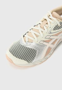 Shoe de corrida com um cabedal em malha em bege e detalhes em prata, apresentando uma sola texturizada e atacadores brancos para um ajuste seguro.