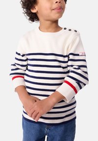 Hvit strikkegenser med marineblå og røde striper, rund halslinning, tre dekorative knapper på skulderen, med ribbestrikkede mansjetter.