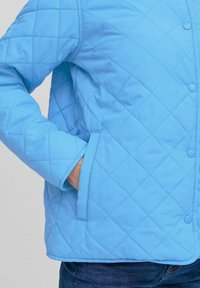 Jachetă cu umplutură de culoare albastru deschis, cu un model în diamant, cu nasturi tip snap și buzunare laterale, având o textură netedă și un design casual.