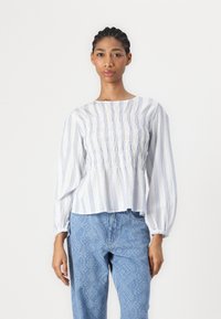 Hvit bluse med blå vertikale striper, smocket liv, og lange puffermer, kombinert med lyseblå mønstrede jeans.